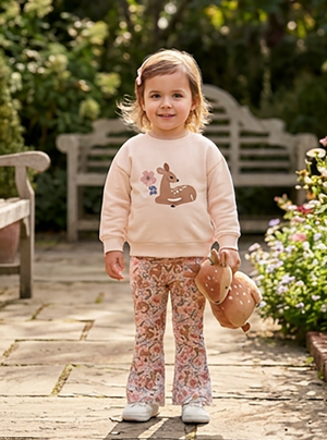 A young child stands on a stone path in a garden wearing a light pink sweatshirt with a deer graphic and matching patterned pants, holding a plush toy.