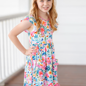 Young girl wearing a colorful floral dress with a pink bow in her hair, standing outdoors.