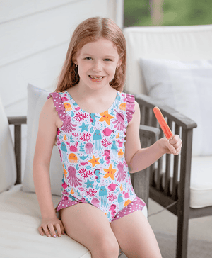 Young girl wearing a pink sea animal themed swimsuit sitting on a chair eating a popsicle, lifestyle, frontMillieloveslily.com