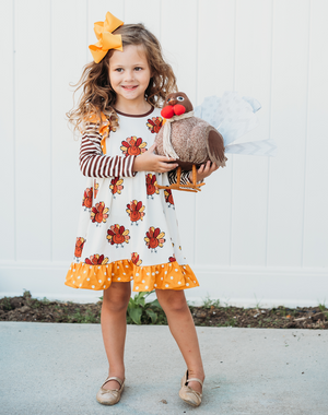 A young girl wearing a cream ground turkey themed printed dress and holding a turkey stuffed animal, lifestyle, Millie Loves Lily
