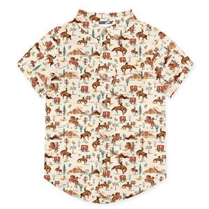 A kids short-sleeve camp shirt with a western-themed all-over print featuring horses, riders, mountains, cacti, and vintage wagons on an off-white background.