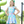 A young girl stands on a playground wearing a light blue sleeveless dress with a colorful bow print. The dress has ruffled shoulders and a ruffled hem, lifestyle