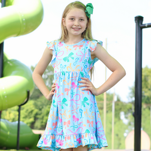 A young girl stands on a playground wearing a light blue sleeveless dress with a colorful bow print. The dress has ruffled shoulders and a ruffled hem, lifestyle