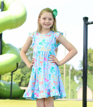 A young girl stands on a playground wearing a light blue sleeveless dress with a colorful bow print. The dress has ruffled shoulders and a ruffled hem, lifestyle
