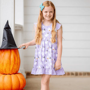 A young girl wearing a lilac halloween themed printed dress standing on a pocrh beside stacked pumpkins, lifestyle, Millie Loves lily