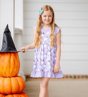 A young girl wearing a lilac halloween themed printed dress standing on a pocrh beside stacked pumpkins, lifestyle, Millie Loves lily