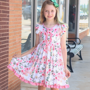 A young girl with blonde hair and a green bow stands outdoors in front of a brick building, wearing a white short-sleeve dress with a pink and green floral print and ruffled trim.
