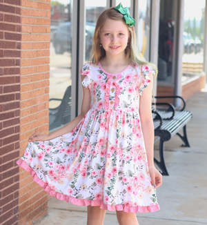 A young girl with blonde hair and a green bow stands outdoors in front of a brick building, wearing a white short-sleeve dress with a pink and green floral print and ruffled trim.