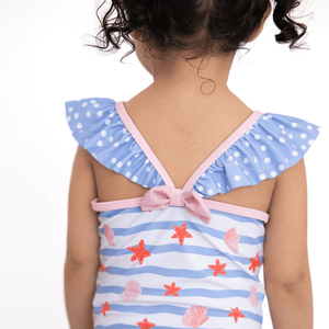 A young girl wearing a blue stripe 1-pc swimsuit with seashells and starfish and blue dot ruffles and a pink back bow, backview, lifestyle