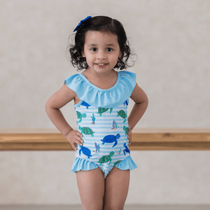 A young girl wearing a blue turtle patterned swimsuit with ruffles standing inside with her hands on her hips, lifestyle