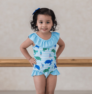 A young girl wearing a blue turtle patterned swimsuit with ruffles standing inside with her hands on her hips, lifestyle