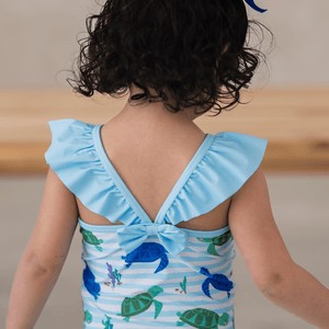 A young girl wearing a blue turtle patterned swimsuit  with shoulder ruffles and a back bow, back 