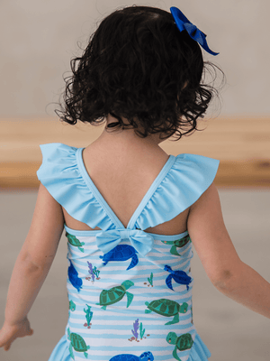 A young girl wearing a blue turtle patterned swimsuit  with shoulder ruffles and a back bow, back 