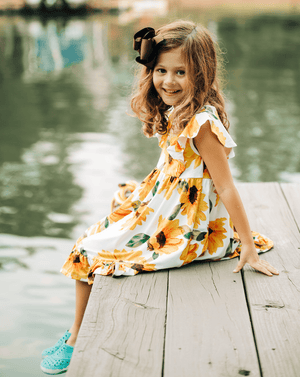Young girls wearing a yellow  & Cream Sunflower dress  sitting on a dock, lifestyle, side- Millieloveslily.com