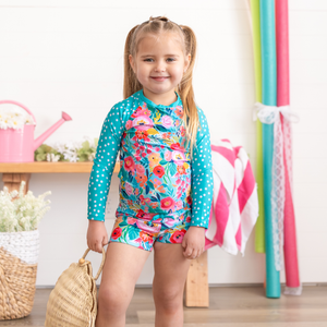 A young  girl wearing a long sleeve turquoise floral and dot rashguard set  holding a straw purse standing in a pool room, lifestyle- Millieloveslily.com