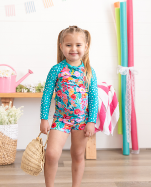 A young  girl wearing a long sleeve turquoise floral and dot rashguard set  holding a straw purse standing in a pool room, lifestyle- Millieloveslily.com