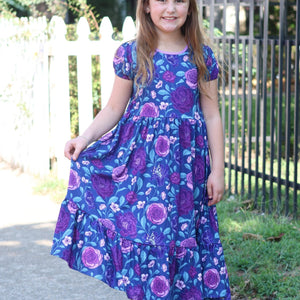 A young girl wearing a  Navy and Purple Floral Hi - Low Midi Dress with short sleeves standing outside in front of a fence, lifestyle