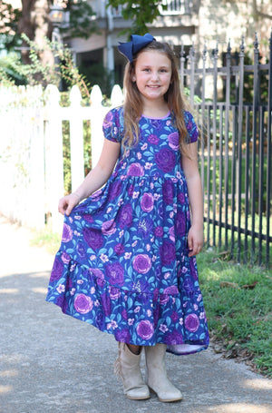 A young girl wearing a  Navy and Purple Floral Hi - Low Midi Dress with short sleeves standing outside in front of a fence, lifestyle