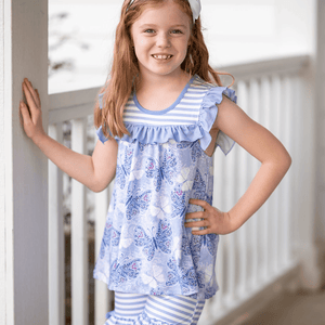 A young girl standing on a porch wearing a purple butterfly themed top and matching stripe short set, lifestyle