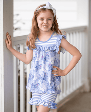 A young girl standing on a porch wearing a purple butterfly themed top and matching stripe short set, lifestyle