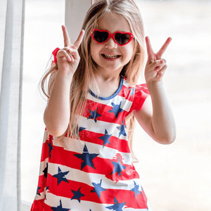 A young girl wearing a patriotic themed stars and stripes top and navy dot shorts holding up peace sign fingers, lifestyle