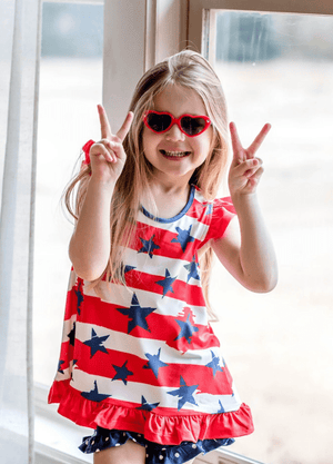 A young girl wearing a patriotic themed stars and stripes top and navy dot shorts holding up peace sign fingers, lifestyle