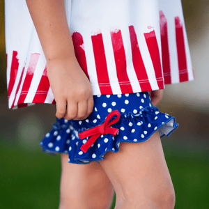 A closeup of a young girl standing outside wearing a matching red, white & blue stars and stripe short set with her hand on her hip, lifestyle