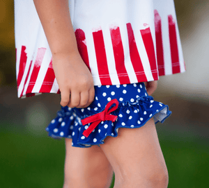 A closeup of a young girl standing outside wearing a matching red, white & blue stars and stripe short set with her hand on her hip, lifestyle