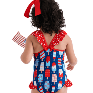A young girl wearing a red,white & blue pospsicle print 1-pc swimsuit with red dot ruffles and and red bow, back