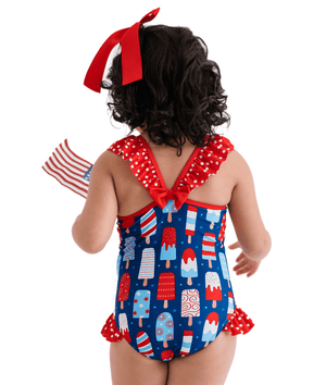A young girl wearing a red,white & blue pospsicle print 1-pc swimsuit with red dot ruffles and and red bow, back