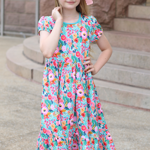 A young girl wearing a turquoise floral hi-low mid-length dress with short sleeves standing in front of steps outside, lifestyle
