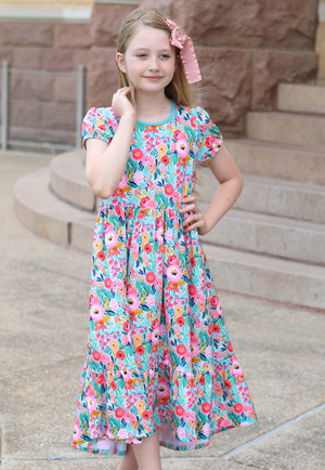 A young girl wearing a turquoise floral hi-low mid-length dress with short sleeves standing in front of steps outside, lifestyle