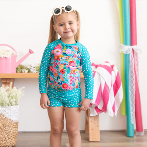 A young girl wearing a turquoise dot and Floral Rashguard Set and white floral sunglasses standing in a pool room, lifestyle- Millieloveslily.com