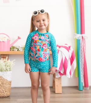A young girl wearing a turquoise dot and Floral Rashguard Set and white floral sunglasses standing in a pool room, lifestyle- Millieloveslily.com