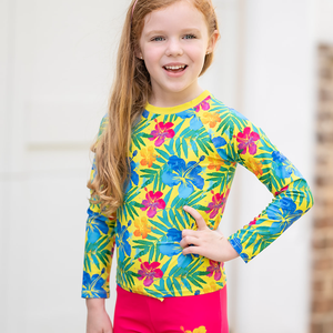 A young girl wearing a yellow & fuschia  Hawaiian tropical pattern Long Sleeve Rashguard and swim short Set, standing in front of a white background, lifestyle UPF 50+, 