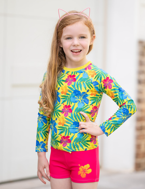 A young girl wearing a yellow & fuschia  Hawaiian tropical pattern Long Sleeve Rashguard and swim short Set, standing in front of a white background, lifestyle UPF 50+, 