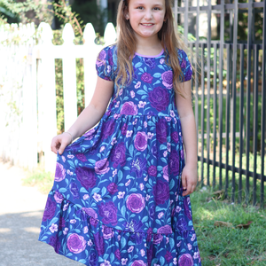 A young girl wearing a  Navy and Purple Floral Hi - Low Midi Dress with short sleeves standing outside in front of a fence, lifestyle