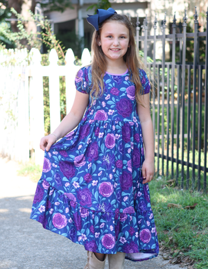 A young girl wearing a  Navy and Purple Floral Hi - Low Midi Dress with short sleeves standing outside in front of a fence, lifestyle