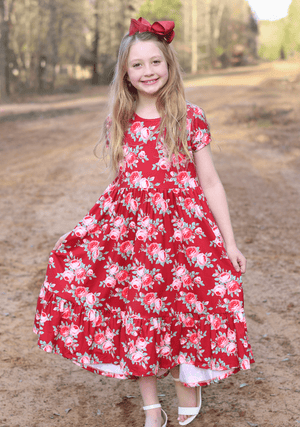 A young girl wearing a red floral hi-low mid-length dress standing outside is a forest like area, lifestyle