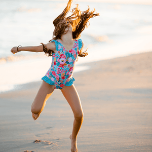 Young girl wearing a floral swimsuit jumping in the air on the beach, lifestyle, front - Millieloveslily.com