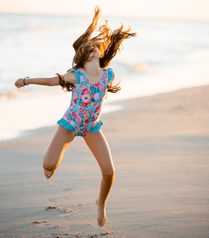 Young girl wearing a floral swimsuit jumping in the air on the beach, lifestyle, front - Millieloveslily.com