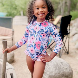 A young girl wearing a Coral flower Ruffle - Accent One - Piece Rashguard standing on the beach, lifestyle, front - Millieloveslily.com