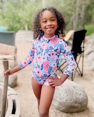 A young girl wearing a Coral flower Ruffle - Accent One - Piece Rashguard standing on the beach, lifestyle, front - Millieloveslily.com