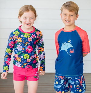 A young girl and boy wearng rashguard sets standing in front of a white wall, lifestyle