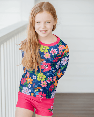 A young girl wearing a  navy and pink  Floral Long Sleeve Rashguard and swim shorts Set standing on a porch, lifestyle- 