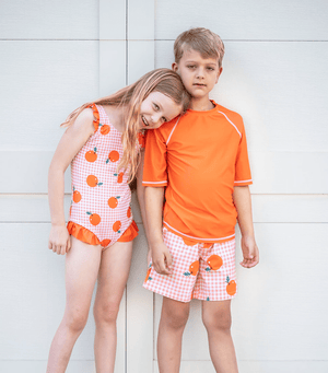 A young girl and boy wearing matching orange gingham swimsuits standing in front of a white wall outside, front Millieloveslily.com
