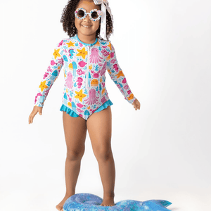 Young girl wearing a white  & turquoise Ocean themed Ruffle - Accent One - Piece Rashguard and white flower sunglasses on a white background, front Millieloveslily.com