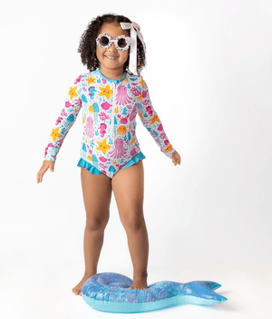 Young girl wearing a white  & turquoise Ocean themed Ruffle - Accent One - Piece Rashguard and white flower sunglasses on a white background, front Millieloveslily.com