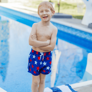 Boy wearing  Red, Wht, & Blue  stars Swim Shorts standing in front of a pool, front