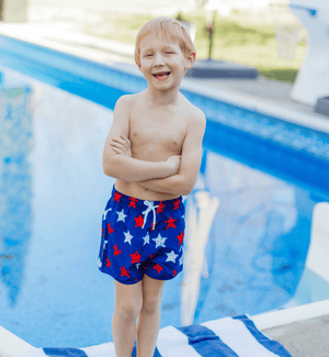 Boy wearing  Red, Wht, & Blue  stars Swim Shorts standing in front of a pool, front